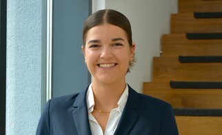 Portrait photo of Lilian Vogt, Executive Assistant, wearing a dark blue blazer and white blouse, standing in front of a staircase and smiling at the camera